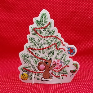 May include: A white felt Christmas tree with a red and green embroidered design. A small mouse is holding a pink ornament. The tree is decorated with red beads and ornaments.