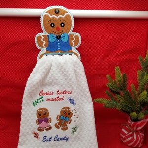 May include: White kitchen towel with embroidered gingerbread people and the text "Cookie testers NOT wanted Eat Candy". A gingerbread person towel holder is hanging on a white bar.