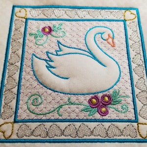 Pode incluir: Cisne branco bordado com detalhes florais em turquesa, rosa e dourado. O cisne é rodeado por uma moldura turquesa com uma borda de corações prateados e dourados.
