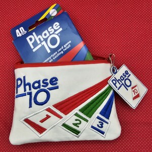 May include: A white canvas zippered pouch with the words "Phase 10" embroidered on the front. The pouch has a red, green, and blue striped design with the numbers 1, 2, and 3 embroidered on the stripes. A small red and white tag with the words "Phase 10" and the number 10 is attached to the zipper pull. A blue and white box with the words "Phase 10" is sitting on top of the pouch.