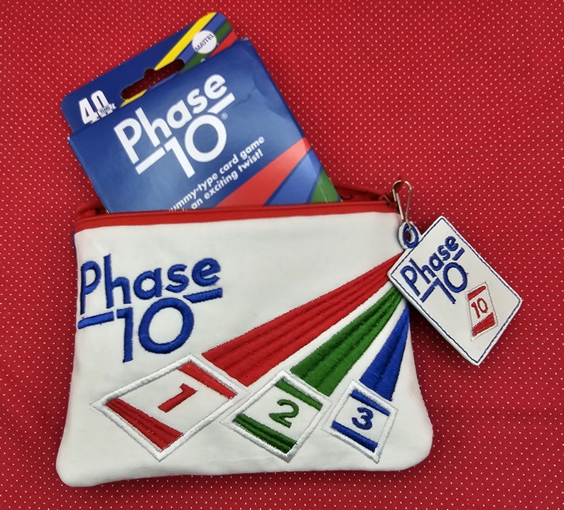 May include: A white canvas zippered pouch with the words "Phase 10" embroidered on the front. The pouch has a red, green, and blue striped design with the numbers 1, 2, and 3 embroidered on the stripes. A small red and white tag with the words "Phase 10" and the number 10 is attached to the zipper pull. A blue and white box with the words "Phase 10" is sitting on top of the pouch.