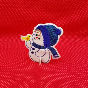 May include: Embroidered white snowman with a blue hat and scarf holding a yellow bird. The snowman is on a red background with white polka dots.