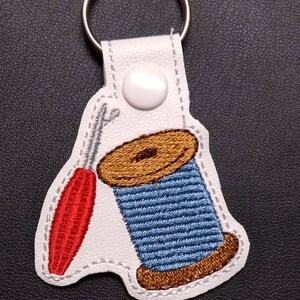 以下が含まれることがあります： 赤い針と青と茶色の糸巻きが刺繍された白い革のキーホルダーです。