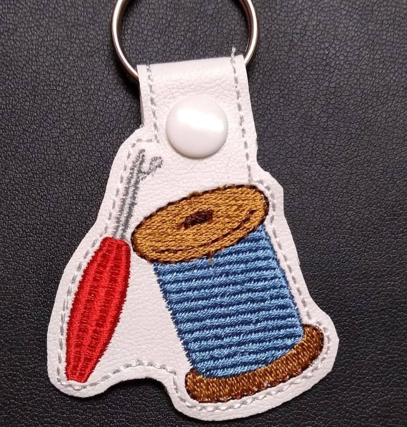以下が含まれることがあります： 赤い針と青と茶色の糸巻きが刺繍された白い革のキーホルダーです。