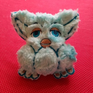 May include: A plush toy in the shape of a small creature with large, expressive blue eyes and a light blue, fluffy body. The toy has embroidered details in teal and brown, with a beak-like feature and small feet.