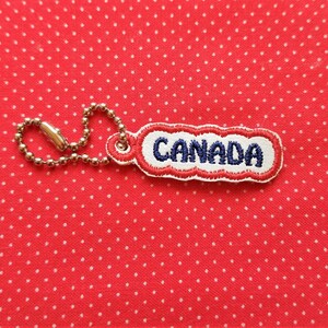 Puede incluir: Un llavero de tela rojo, blanco y azul con la palabra "CANADA" bordada en hilo azul.