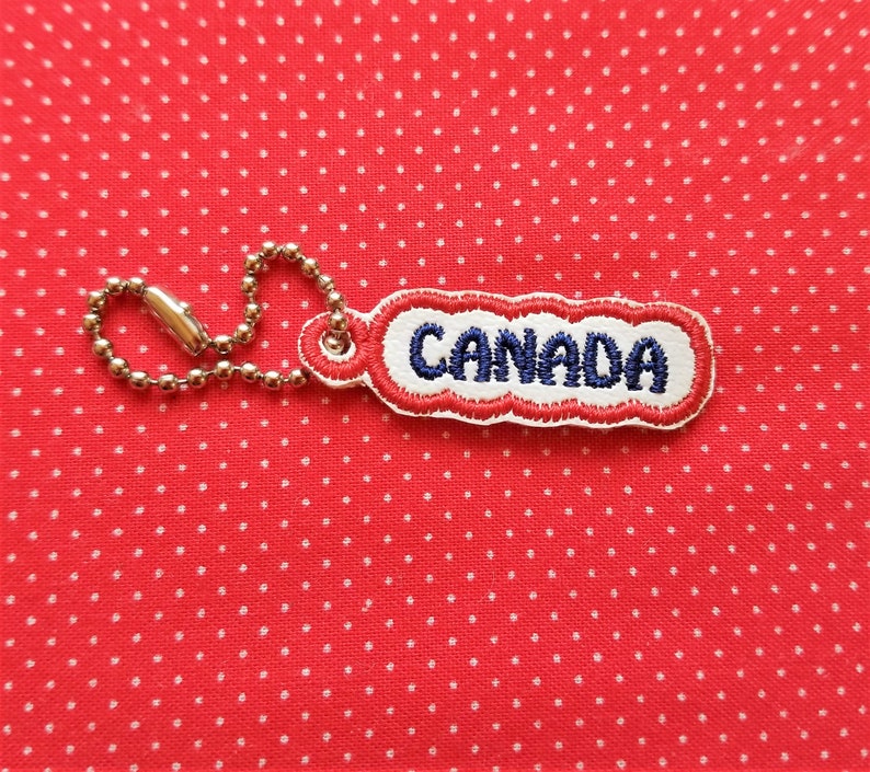 Puede incluir: Un llavero de tela rojo, blanco y azul con la palabra "CANADA" bordada en hilo azul.