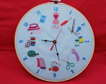 Puede incluir: Un reloj de aro de bordado azul y blanco con imágenes de temática de costura, que incluyen una máquina de coser, carretes de hilo, tijeras y un alfiletero. La esfera del reloj tiene números romanos y una aguja negra y roja.