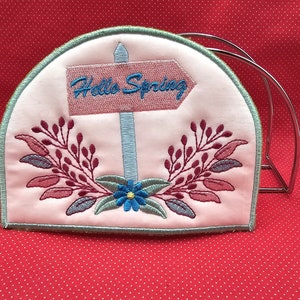 May include: A pink fabric tea cozy with embroidered floral designs and a sign that reads "Hello Spring".