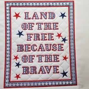 May include: A white quilted fabric square with a red, white, and blue embroidered border. The center of the square features the text "LAND OF THE FREE BECAUSE OF THE BRAVE" in red and blue embroidered letters.
