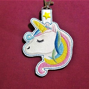 May include: Embroidered unicorn keychain with a white face, a yellow horn, and a rainbow mane. The keychain has a white strap with a yellow star and a silver clasp. The unicorn is outlined in blue stitching.