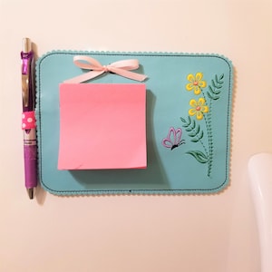 Puede incluir: Un tablero de notas de fieltro azul claro con un borde festoneado. El tablero tiene una nota adhesiva rosa con un lazo de cinta blanco y un diseño bordado de flores amarillas y una mariposa morada.