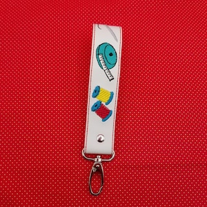 May include: White leather key chain with a blue measuring tape, yellow and red spools of thread, and a silver key ring.