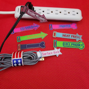 Pode incluir: Um conjunto de organizadores de cabos coloridos com etiquetas bordadas para diferentes dispositivos eletrônicos, incluindo um modem de roteador, uma unidade de CD/DVD, um apontador de lápis, uma impressora, uma prensa térmica, um telefone celular e uma base de superfície. Os organizadores estão sobre um fundo vermelho.