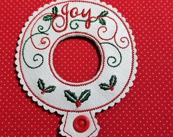 May include: White felt Christmas ornament with red and green embroidery. The ornament is shaped like a wreath with the word "Joy" in red cursive lettering. There is a red button on the bottom of the ornament.