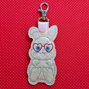 May include: A white bunny keychain with pink ears and heart-shaped glasses. The bunny is wearing blue and red glasses with a pink nose and mouth. The keychain has a silver loop.