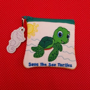 May include: A small white zippered pouch with a green sea turtle embroidered on the front. The pouch has a yellow sun and blue waves embroidered around the turtle. The text "Save the Sea Turtles" is embroidered in yellow at the bottom of the pouch.