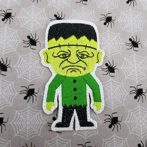 May include: Green Frankenstein monster character with black hair, green shirt, and black pants. The character is embroidered on a white fabric with a spiderweb pattern.