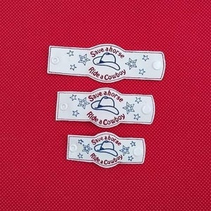 May include: Three white fabric wristbands with blue embroidery. Each wristband features a cowboy hat and the text "Save a horse, Ride a Cowboy".