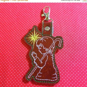 May include: A brown felt Christmas ornament with a white outline of a kneeling shepherd boy holding a staff. The shepherd is looking up at a yellow star with white rays. The ornament has a silver metal key ring attached.