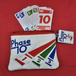 Könnte beinhalten: Eine weiße Canvas-Tasche mit Reißverschluss, auf der die Worte "Phase 10" in Blau gestickt sind. Die Tasche hat ein rot-grün-blaues Streifenmuster. Ein kleiner weißer Anhänger mit den Worten "Phase 10" in Blau ist am Reißverschluss befestigt. Im Hintergrund ist ein Stapel Spielkarten mit der Nummer "10" zu sehen.