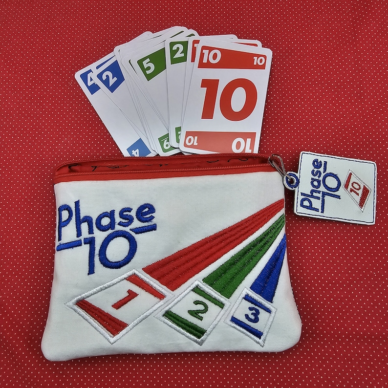 May include: White canvas zippered pouch with the words "Phase 10" embroidered in blue. The pouch has a red, green, and blue striped design. A small white tag with the words "Phase 10" in blue is attached to the zipper pull. A stack of playing cards with the number "10" on them is visible in the background.