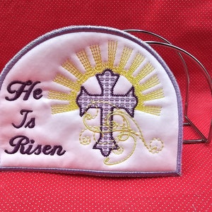 May include: White fabric napkin ring with embroidered purple cross, yellow sun rays, and the text "He Is Risen".