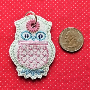 Può includere: Portachiavi in ​​cuoio bianco a forma di gufo con ricami rosa e blu. Il gufo ha un fiore sulla testa e un motivo a scacchi rosa e bianco sul petto. Il portachiavi è circa delle dimensioni di una moneta da 25 centesimi di dollaro americano.