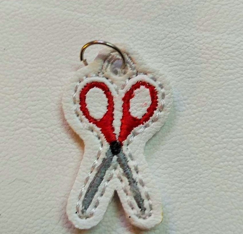 Puede incluir: Un llavero de cuero blanco con un par de tijeras bordadas en rojo y plata.