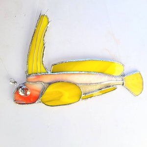 May include: A stained glass fish with a yellow body, pink and red head, and silver accents. The fish is hanging from a string.