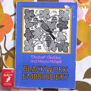 May include: A vintage book titled "Blackwork Embroidery" by Elisabeth Geddes and Moyra McNeill. The cover is blue with a black and white illustration of a bird and grapevines. A small red matchbox with the text "Redheads" is next to the book.