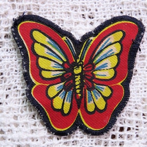 May include: A vibrant butterfly patch with a red body and wings, edged in navy blue. The wings display yellow, blue, and red patterns. The patch is probably fabric and is set against a white, textured background.