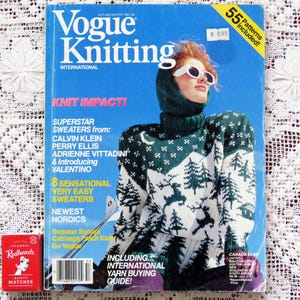 May include: A vintage Vogue Knitting International magazine with a blue cover featuring a woman in a green and white sweater. The magazine includes text such as "Knit Impact!" and "55 Patterns Included!" A red matchbox is in the lower left corner.