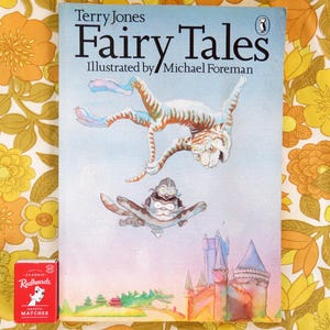 May include: A paperback book titled Terry Jones Fairy Tales, illustrated by Michael Foreman, featuring a tiger and a small creature flying above a castle. Next to the book is a small red box of Redheads brand safety matches.