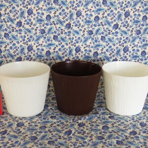 May include: Three plastic plant pots, one white, one brown, and one white. The pots are lined up in a row on a patterned fabric background.