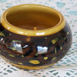 May include: A small, brown ceramic bowl with a yellow glaze on the inside and a geometric pattern on the outside. The bowl is sitting on a white crocheted surface.