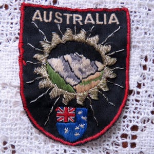May include: Embroidered patch with the word "AUSTRALIA" at the top. The patch features a mountain scene within a gold sunburst design. A small blue shield with the Australian flag and Southern Cross stars is at the bottom. The patch has a red border.