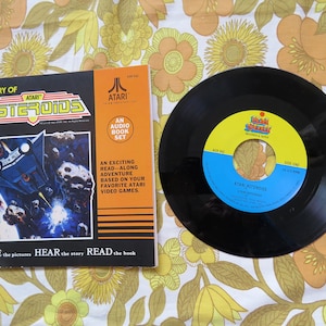 May include: A black vinyl record with a blue and white label that says "Atari Asteroids". The record is on a white and yellow floral tablecloth next to a book titled "Story of Atari Asteroids". The book is an audio book set with a picture of a spaceship on the cover.
