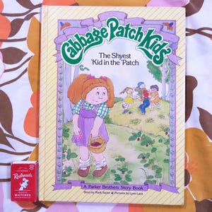 May include: A vintage "Cabbage Patch Kids" storybook, "The Shyest 'Kid in the 'Patch," featuring a cartoon illustration of a girl in a purple dress. A box of "Redheads" matches is next to the book. The book has a yellow cover.