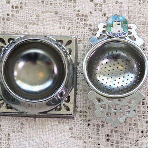 May include: Two silver tea strainers. One is square with a floral design, and the other is round with a decorative handle featuring a lighthouse image. Both are made of metal and are designed for steeping loose leaf tea.