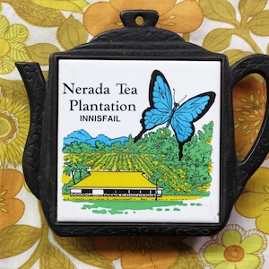 May include: Black cast iron trivet shaped like a teapot. It has a white tile insert with a blue butterfly and a landscape scene. The text on the tile reads "Nerada Tea Plantation INNISFAIL".