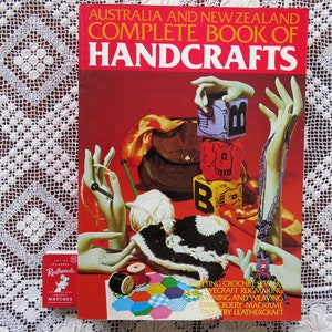 May include: A book titled "Australia and New Zealand Complete Book of Handcrafts" with a red cover. The cover features illustrations of crafting tools and projects, including knitting, crochet, and weaving. A box of Redheads matches is next to the book.