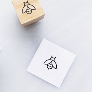 May include: A wooden stamp with a black bee design and a stamped image of the same bee on a white square. The stamp is rectangular and made of light-colored wood. The bee design is simple and outlined in black.