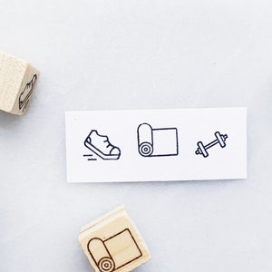 May include: Four wooden stamps with different fitness icons: a running shoe, a yoga mat, a dumbbell, and a yoga mat. The stamps are on a white surface.