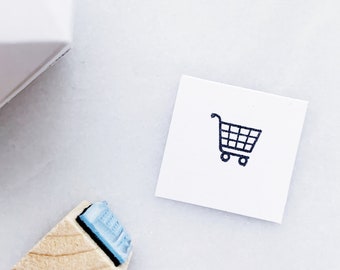 May include: A wooden stamp with a blue rubber insert depicting a shopping cart icon. The stamp is on a white surface next to a white card with a black shopping cart icon.