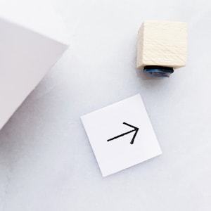 May include: A white wooden block with a blue ink pad on the bottom. The block is used to stamp an arrow pointing to the right on a white piece of paper.