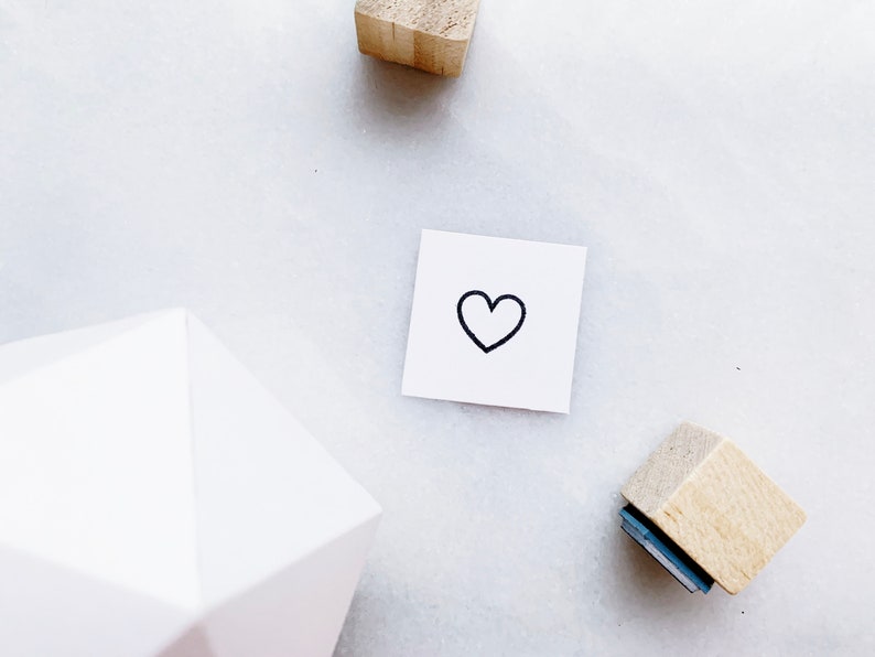 Puede incluir: Una tarjeta blanca con un coraz&oacute;n negro dibujado a mano. La tarjeta est&aacute; sobre una superficie blanca con dos bloques de madera.