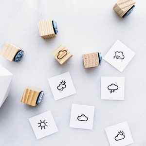 May include: Wooden stamps with weather icons, including a sun, a cloud, a cloud with rain, a cloud with lightning, and a cloud with wind. The stamps are on a white surface with white paper squares with the same icons.