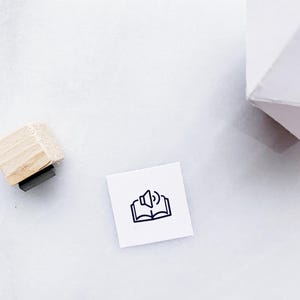 May include: A square rubber stamp with a wooden handle featuring a black line art icon of an open book with a speaker symbol above it, indicating an audiobook or read-aloud function, positioned next to a white geometric object.