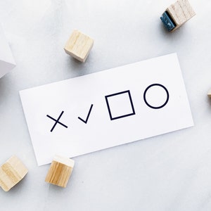 May include: A white card with a checkmark, an X, a square, and a circle. The card is on a white surface with small wooden blocks.